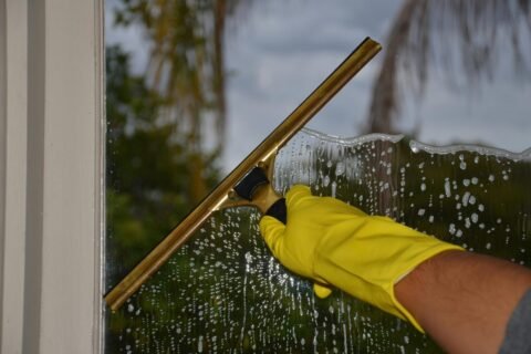 Close-up of hand cleaning window with squeegee outdoors using yellow gloves.
